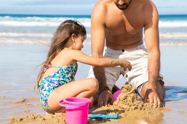 Doce menina e o pai construindo castelo de areia na praia, sentados na areia molhada, curtindo as férias no mar