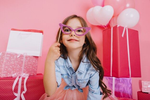 Doce menina retrato com longos cabelos castanhos, segurando a máscara no rosto, olhando para a câmera em caixas de presente, balões, fundo rosa. Criança linda e animada se divertindo, comemorando a festa de aniversário