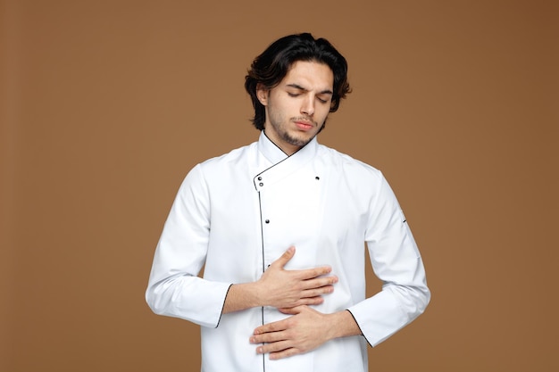 doendo jovem chef masculino vestindo uniforme, mantendo as mãos na barriga com os olhos fechados, isolados no fundo marrom