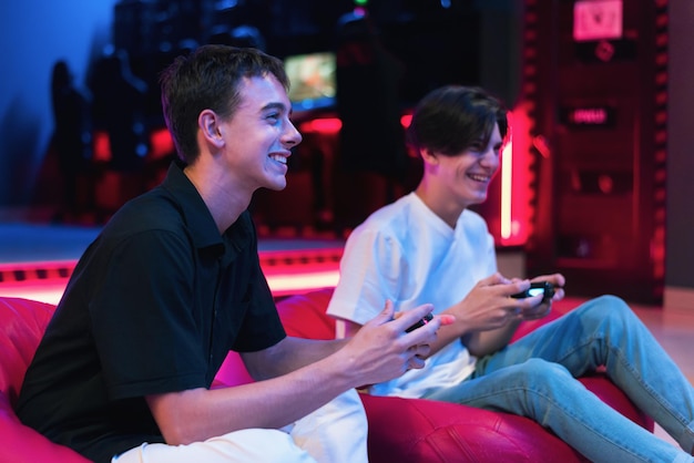 Dois amigos adolescentes estão jogando um console de videogame usando gamepads e sorrindo enquanto estão sentados no feijão