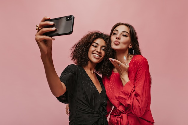 Dois amigos da moda com cabelos escuros em roupas da moda de bolinhas fazendo selfie, sorrindo e mandando beijo na parede rosa