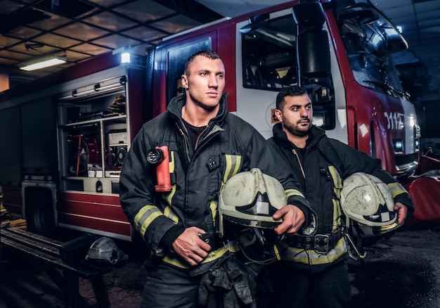 Dois bombeiros vestindo uniforme de proteção ao lado de um caminhão de bombeiros. Chegada de plantão à noite