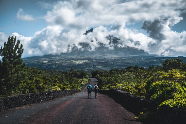 Dois caminhantes andando em uma estrada estreita, rodeada por vegetação com montanha nublada