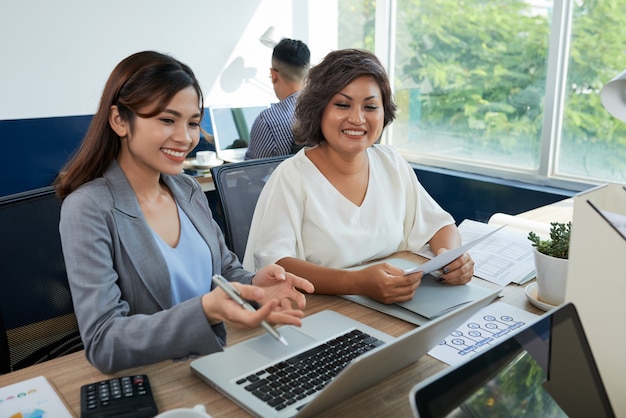 Dois colegas asiáticos estão sentados na mesa no escritório com laptop, uma mulher ajudando outra