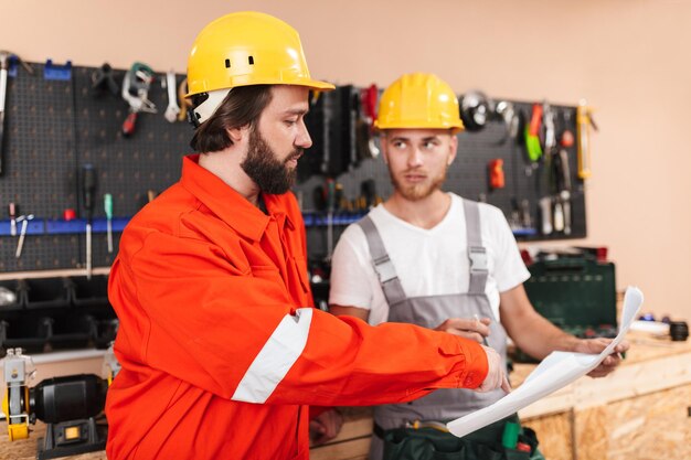 Dois construtores em roupas de trabalho e capacetes amarelos trabalhando em oficina com ferramentas em segundo plano