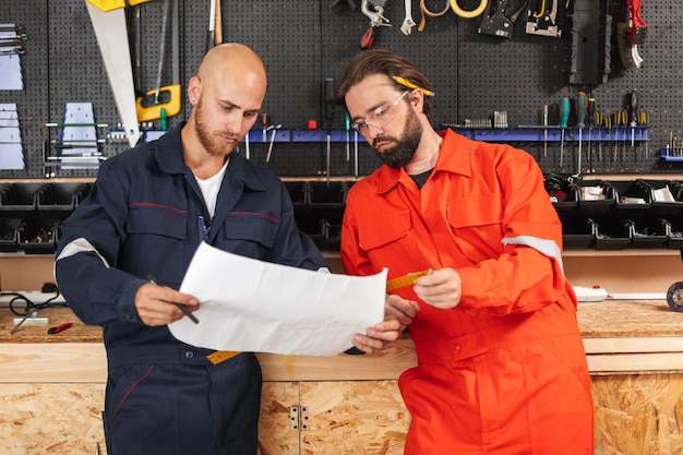 Dois construtores em roupas de trabalho olhando cuidadosamente no plano de esboço com ferramentas em segundo plano na oficina