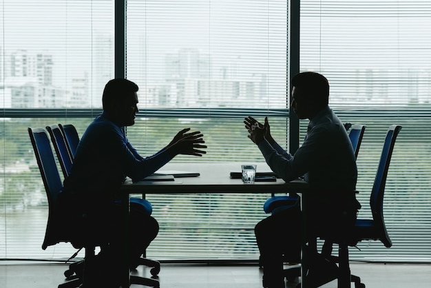 dois contornos humanos contra a janela fechada do escritório, sentados um em frente ao outro e negociando