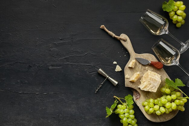 Dois copos de vinho branco e um saboroso prato de queijo com frutas em um prato de madeira na pedra preta