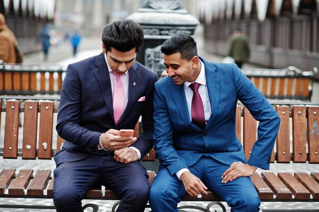 Dois elegantes modelos de homens elegantes indianos de terno posaram no dia de inverno sentados no banco e olhando para o telefone