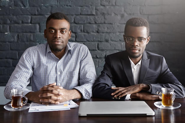 Dois empresários afro-americanos bem-sucedidos bonitos trabalhando no escritório, sentado à mesa com laptop, papéis e canecas