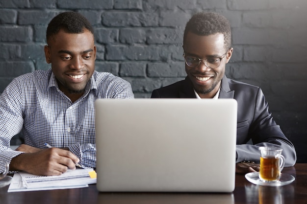 Dois empresários afro-americanos jovens bem sucedidos alegres sentado no interior do escritório moderno na frente do computador portátil aberto, olhando para a tela com um sorriso feliz, discutindo planos de negócios e idéias