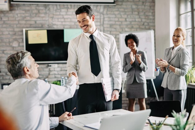 Dois empresários felizes apertando as mãos após um acordo bem-sucedido no escritório enquanto suas colegas as aplaudem O foco está no jovem empresário