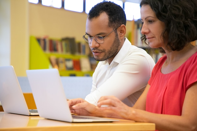 Dois estudantes concentrados conversando e olhando para laptop na biblioteca