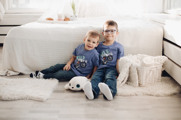 Dois garoto sorridente do sexo masculino posando juntos no interior do confortável quarto branco. irmãos felizes se abraçando, se divertindo na casa aconchegante, sentados no chão perto da cama