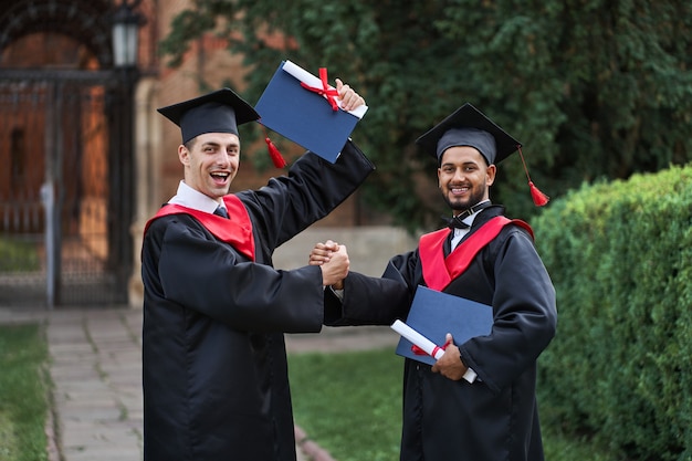 Dois graduados internacionais comemorando a formatura no campus da universidade e olhando para a câmera.