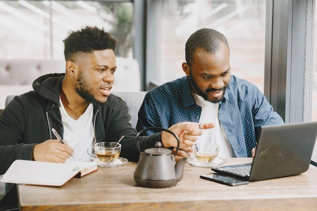 Dois homens afro-americanos trabalhando atrás de um laptop e escrevendo em um caderno. Homens com barba, sentados em um café.