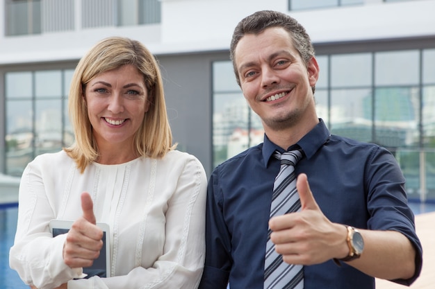 Dois homens felizes e executivos fêmeas que mostram os polegares acima ao ar livre.