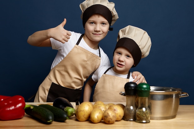 Foto grátis dois irmãos filhos vestindo uniforme de chef preparando o jantar na cozinha: menino confiante mostrando os polegares para cima e abraçando o irmão mais novo, pronto para fazer uma refeição deliciosa com vegetais orgânicos frescos