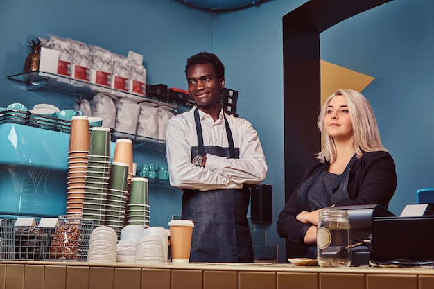 Dois jovens baristas multirraciais em aventais de pé de forma acolhedora em seu café da moda.