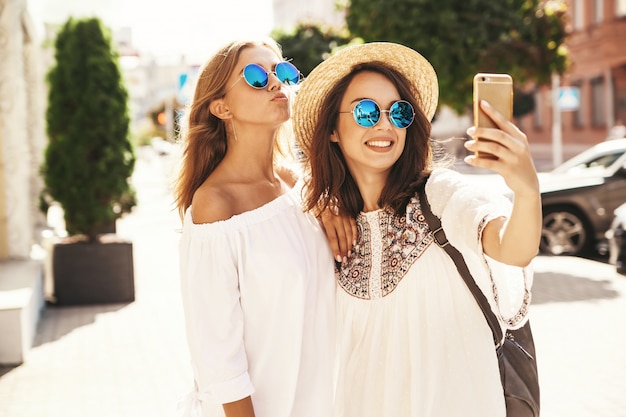 Dois modelos de mulheres jovens loiras e morenas de hippie sorrindo loira no verão hipster branco vestem tirar fotos de selfie para mídias sociais no telefone. Cara de surpresa, emoções,