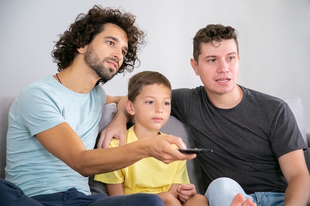 Foto grátis dois pais entusiasmados e filho assistindo programa de tv em casa, sentados no sofá da sala, se abraçando, usando o controle remoto, olhando para longe. conceito de entretenimento familiar e doméstico