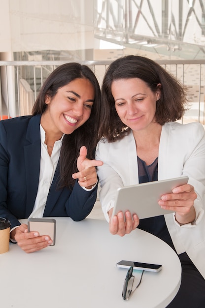 Dois parceiros femininos sorridentes usando gadgets no café moderno