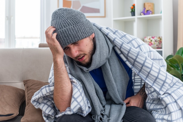 dolorido jovem doente usando cachecol e chapéu de inverno sentado no sofá na sala enrolado em um cobertor, mantendo as mãos na cabeça com os olhos fechados