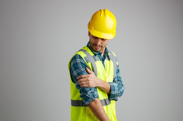 Dolorido jovem engenheiro masculino usando capacete de segurança e uniforme em pé na vista de perfil, mantendo a mão no braço olhando para o braço isolado no fundo branco com espaço de cópia