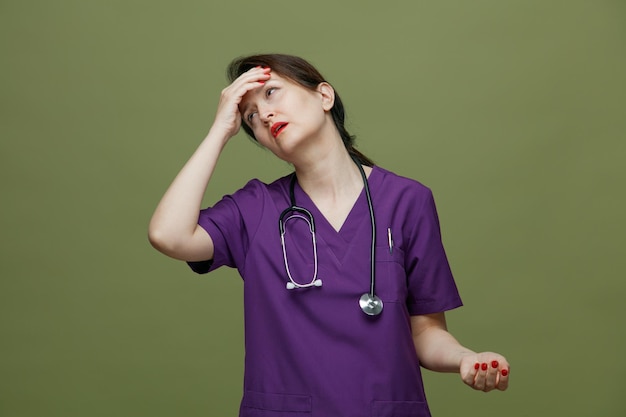 Dolorosa médica de meia-idade vestindo uniforme e estetoscópio no pescoço, mantendo a mão na cabeça, olhando para o lado com dor de cabeça isolada em fundo verde-oliva