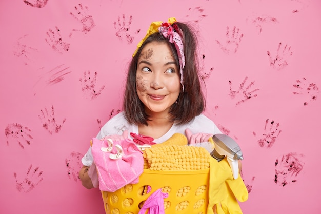 dona de casa com cabelo escuro posa perto de um cesto cheio de roupa suja com o rosto sujo isolado em rosa