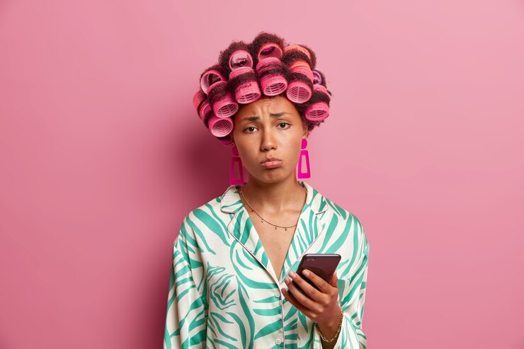 Foto grátis dona de casa descontente namorado triste não liga, segura o celular e aguarda mensagem desejável, chateada por causa do encontro adiado, usa bobes de cabelo e vestido de seda, parece infeliz