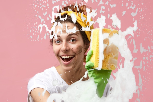 Dona de casa divertida e alegre com manchas sujas no rosto fazendo limpeza de primavera em casa, limpando a espuma densa e espessa do vidro da janela ou do espelho usando pano, detergente e luvas protetoras de borracha