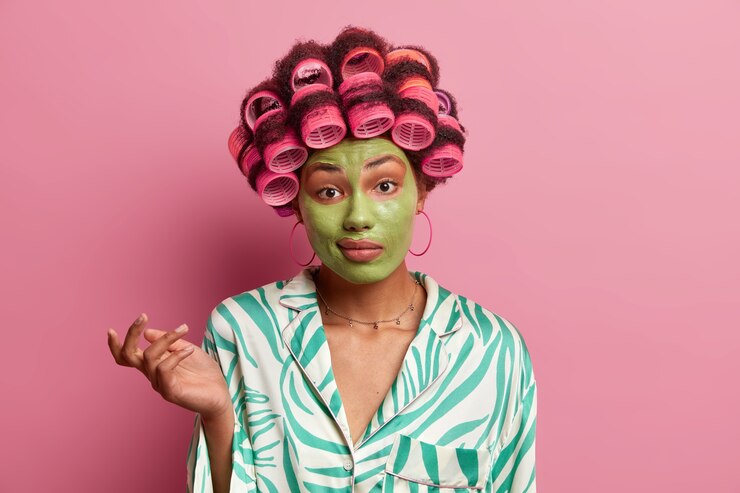 Foto grátis dona de casa duvidosa e perplexa pondera sobre a decisão certa, encolhe os ombros em perplexidade, vestida de pijama, faz penteado com rolos, aplica máscara de beleza verde para refrescar a pele e tez