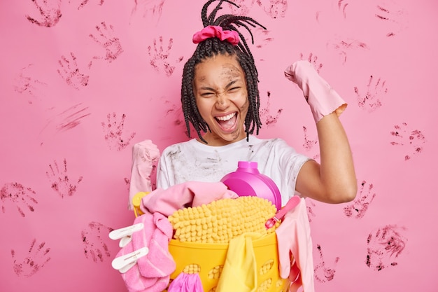 Dona de casa feliz faz gesto de sim com os punhos cerrados celebra o término da limpeza da casa usa luvas de borracha barracas perto do cesto de roupa suja se divertindo isolado sobre a parede rosa suja