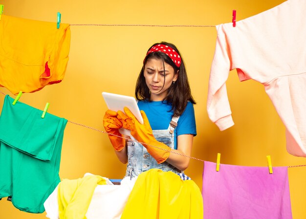 Dona de casa linda e engraçada fazendo trabalhos domésticos isolados no espaço amarelo. Jovem mulher caucasiana rodeada de roupas lavadas