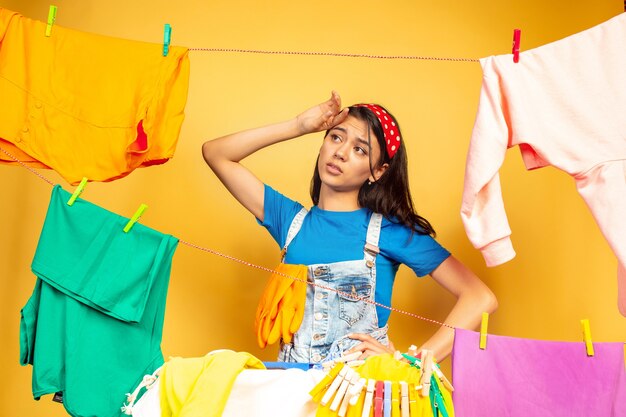 Dona de casa linda e engraçada fazendo trabalhos domésticos isolados no fundo amarelo. jovem mulher caucasiana, rodeada de roupas lavadas. vida doméstica, arte brilhante, conceito de limpeza. triste e cansado.