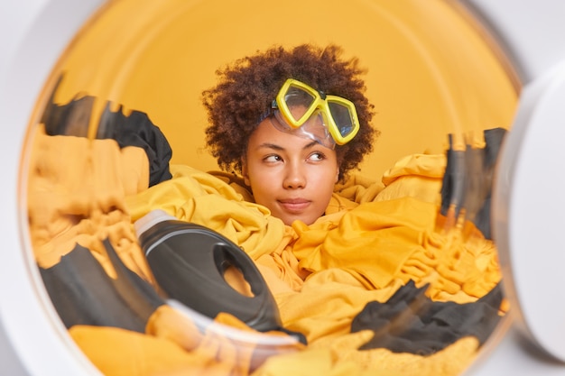 Dona de casa pensativa, de cabelos cacheados, focada e com expressão pensativa, usa máscara de mergulho na testa carrega máquina de lavar com roupa suja faz as tarefas domésticas diárias