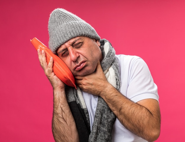 Dorido adulto doente caucasiano homem com lenço no pescoço usando chapéu de inverno coloca a mão no pescoço e segura a garrafa de água quente isolada na parede rosa com espaço de cópia