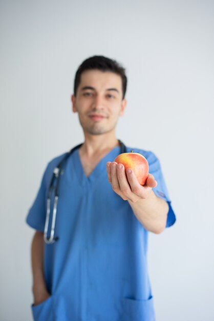 Doutor masculino de sorriso que guarda e que oferece a maçã vermelha com foco na maçã.