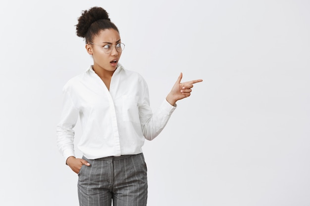 Droga, garota, o que você está vestindo. Retrato de uma colega de trabalho descontente, intensa e confusa, de pele escura, de óculos e terno, apontando e olhando para a direita com expressão de perplexidade e antipatia