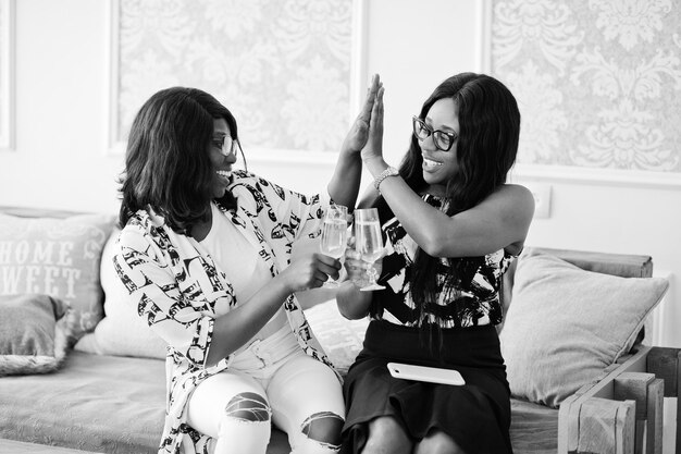 Duas amigas africanas usam óculos posando em sala branca interna e bebendo champanhe