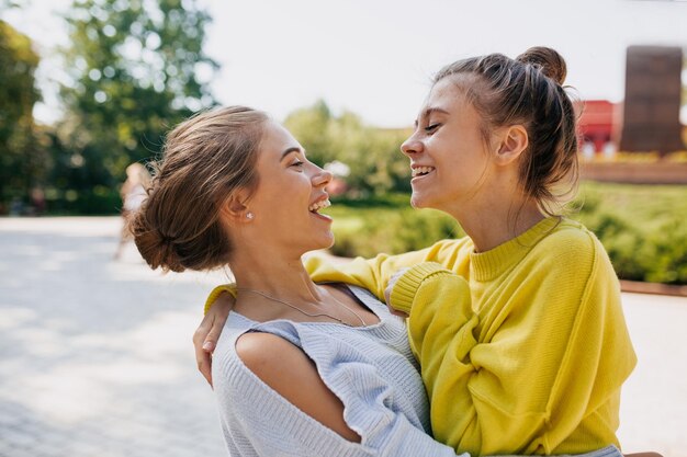 Duas amigas emocionais vestindo suéteres amarelos e azuis estão rindo e abraçando enquanto caminhavam no parque