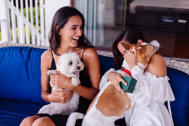 Duas amigas lindas e felizes relaxando em casa no sofá, sorrindo e brincando com cachorros