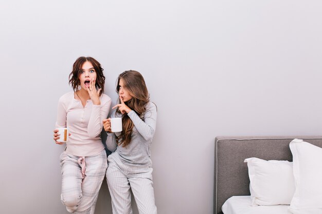 Duas belas garotas de pijama com xícaras, se divertindo no quarto na parede cinza. Menina com cabelo encaracolado parece surpresa.