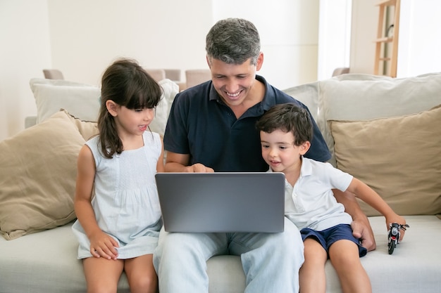 Duas crianças felizes e seu pai usando o laptop enquanto está sentado no sofá em casa, olhando para a tela.
