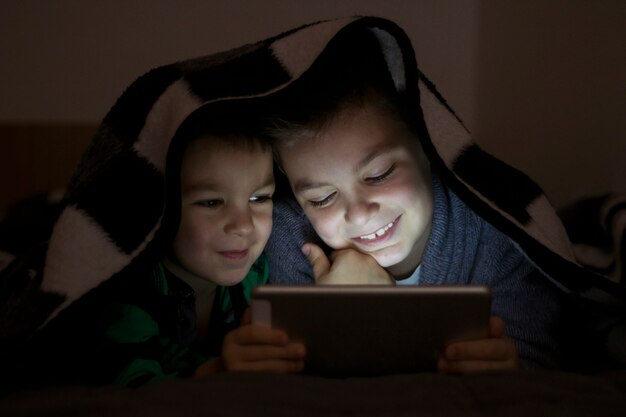 Duas crianças usando tablet pc sob cobertor durante a noite. Irmãos bonitos com tablet computador em um quarto escuro sorrindo.