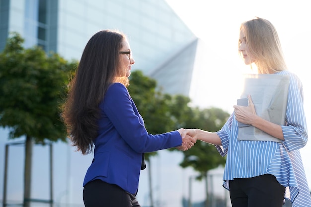 Duas empresárias sérias se cumprimentando com um aperto de mão fora de seus escritórios.