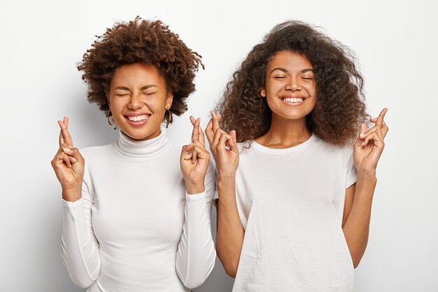 Duas esperançosas mulheres afro-americanas oram, mantêm os dedos cruzados, sorriem amplamente, vestidas com roupas casuais brancas, antecipam resultados positivos, ficam próximas uma da outra.