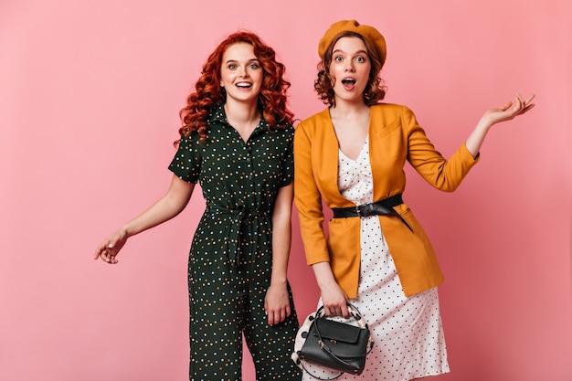 Duas garotas animadas gesticulando enquanto olham para a câmera. Mulher francesa encaracolada posando com um amigo no fundo rosa.