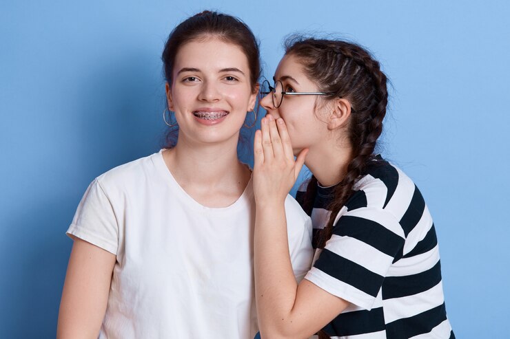 Duas garotas animadas vestidas com roupas de verão fofocando isoladas, senhora de óculos sussurrando algo no ouvido, meninas expressando felicidade.